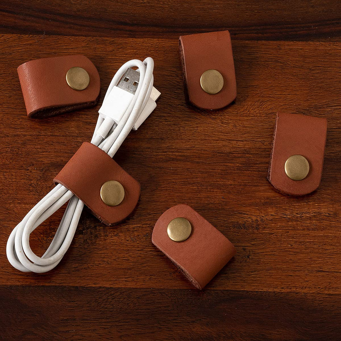A set of 5 brown leather cable holder clips with brass snap buttons, attached to a white USB cable, placed on a wooden surface.
