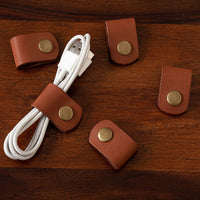 A set of 5 brown leather cable holder clips with brass snap buttons, attached to a white USB cable, placed on a wooden surface.