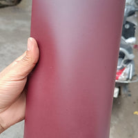 A hand holding a large roll of matte burgundy or dark red material, with an outdoor concrete ground and a scooter visible in the blurred background.