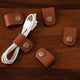 A set of 5 brown leather cable holder clips with brass snap buttons, attached to a white USB cable, placed on a wooden surface.